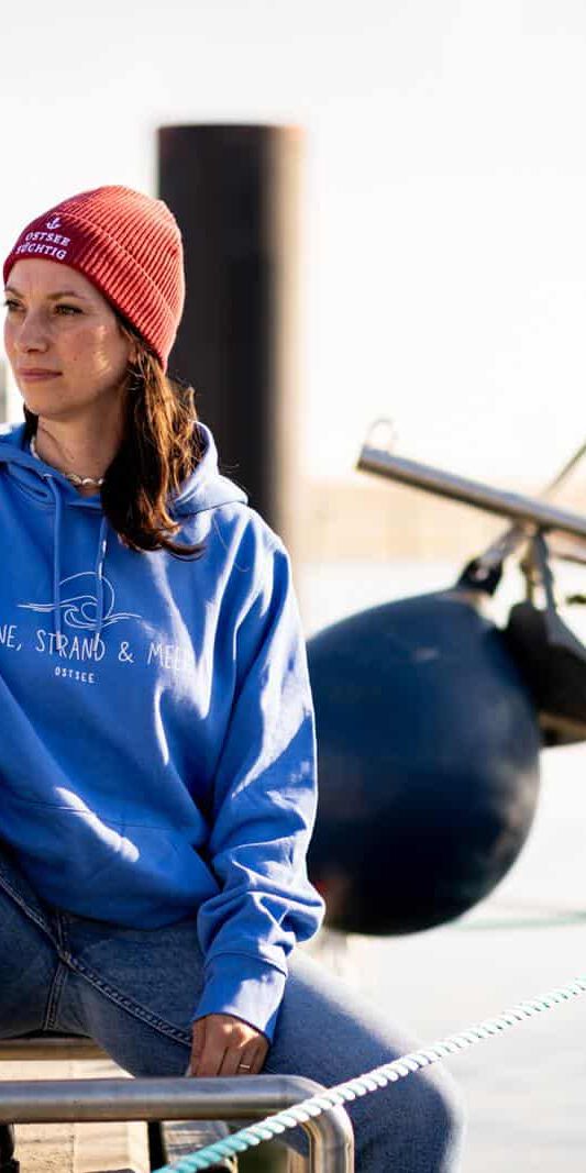 Frau in blauem Hoodie und roter Mütze sitzt mit einer Tasse auf einem Steg im Hafen